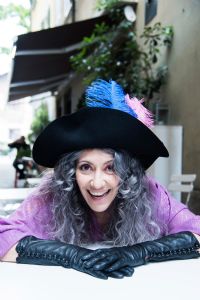 Grey hair model Valeria Sechi smiling and wearing gloves and a hat with feathers