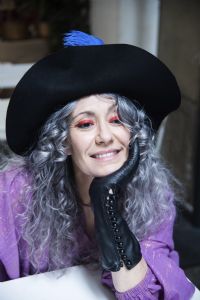 Grey hair model Valeria Sechi smiling and wearing gloves and a hat with feathers